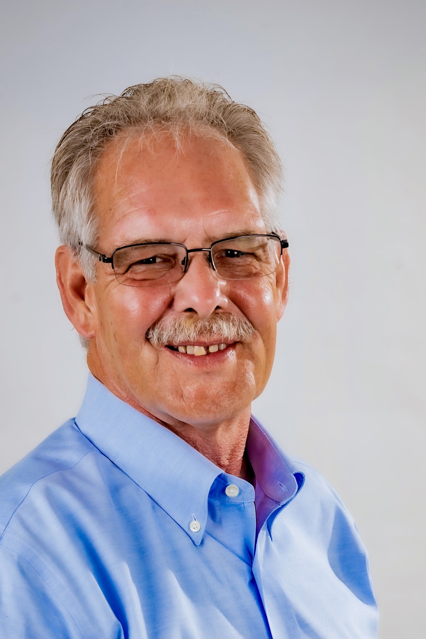 Smiling older man with glasses and a mustache, wearing a light blue collared shirt, against a plain background. Suitable for a professional profile.