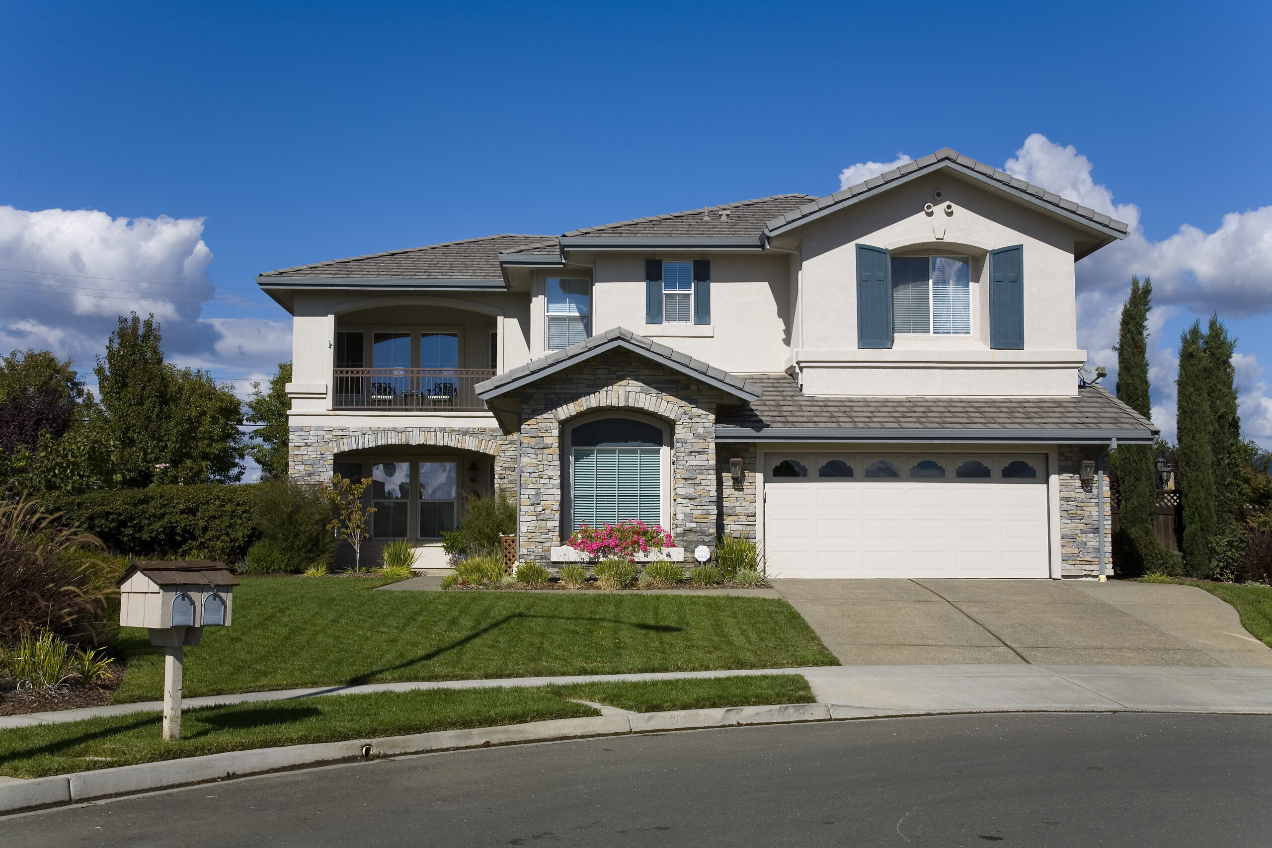 Spacious two-story house featuring stone accents, blue shutters, and a manicured lawn, set against a clear blue sky.