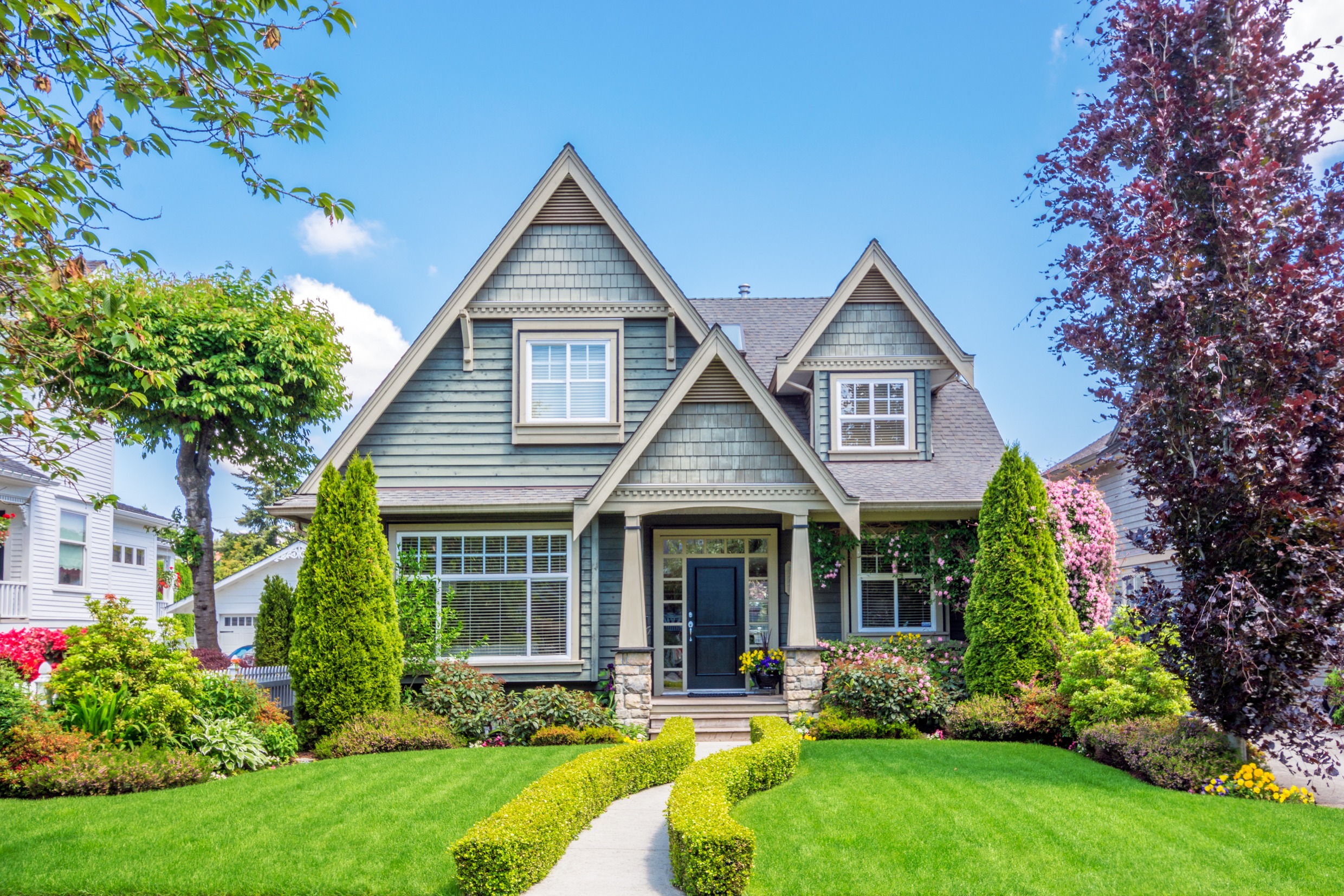 Charming gray cottage-style home with landscaped garden, manicured hedges, and colorful flowers, set against a clear blue sky.